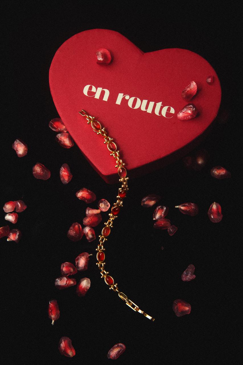 Close up of gold jewelry bracelet with red stones draped over a heart-shaped box and pomegranates.