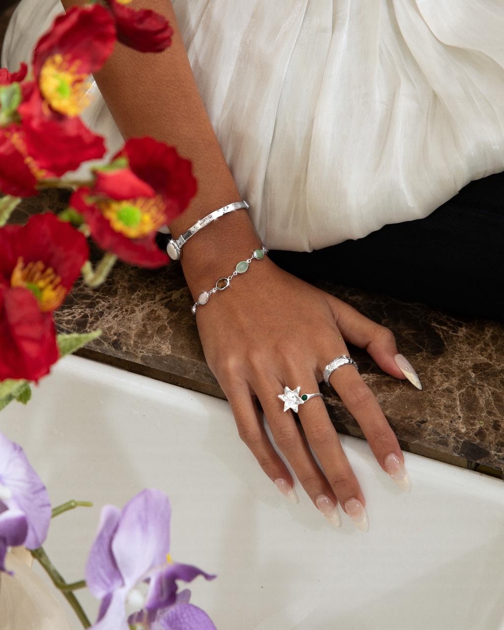 Woman's hand resting near red flowers wearing a silver bangle, a stone chain bracelet, and a large silver star ring.