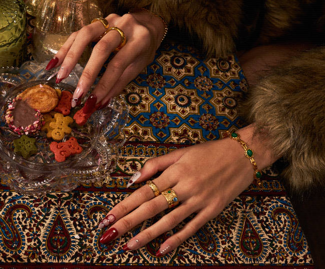 A woman's hands adorned with gold rings and a green gemstone bracelet rest on a patterned cloth