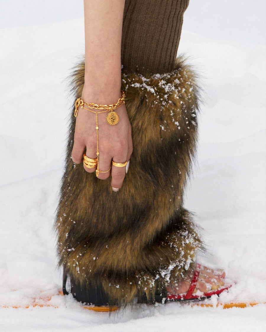 Golden jewelry on a hand resting on a fur-trimmed boot in the snow.