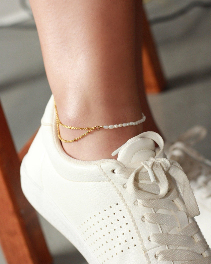 Modern layered anklet featuring a gold chain and delicate white pearls worn with a casual white leather sneaker.