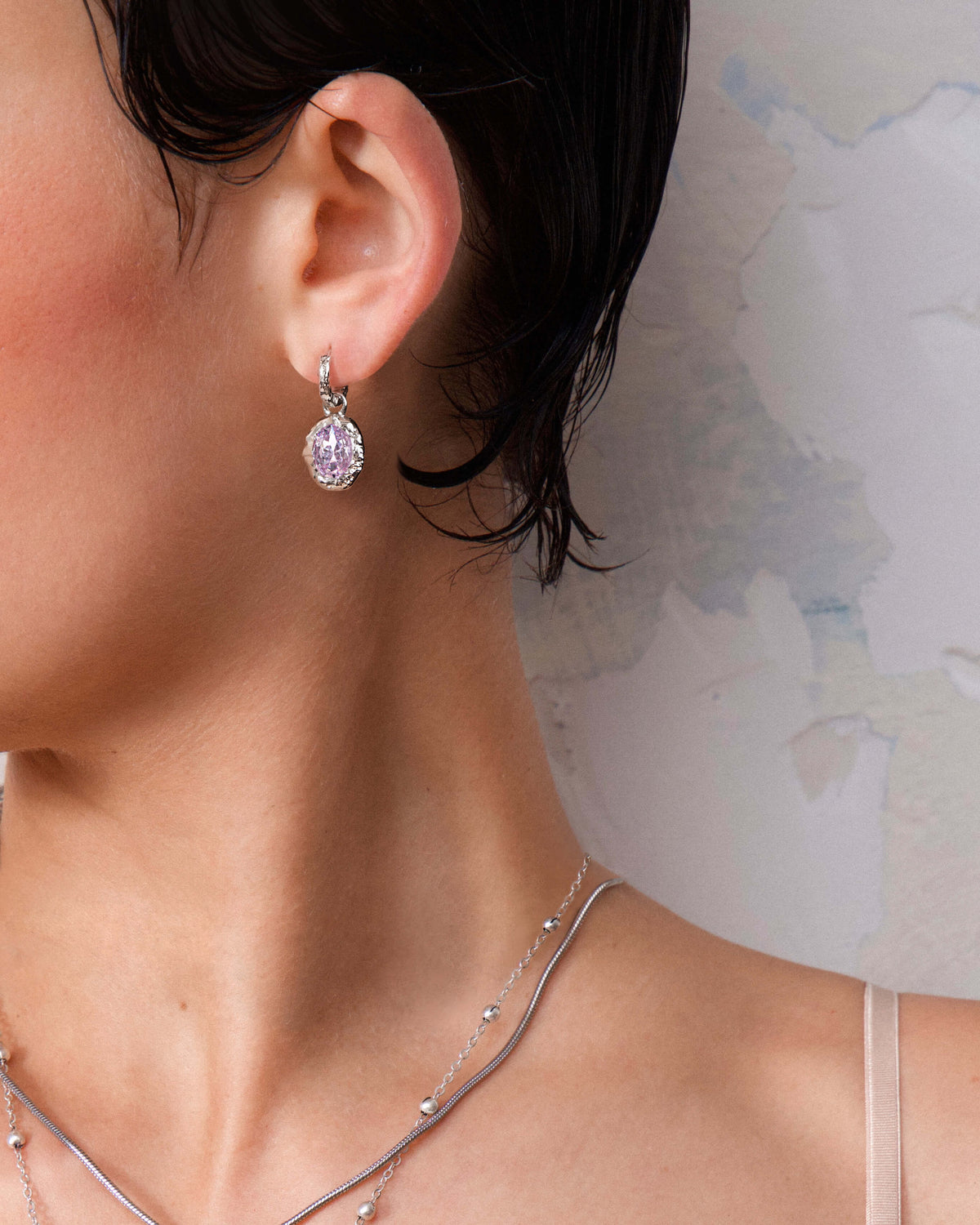 Silver huggie hoop earring with a dangling oval pink gemstone, complementing a silver layered necklace.