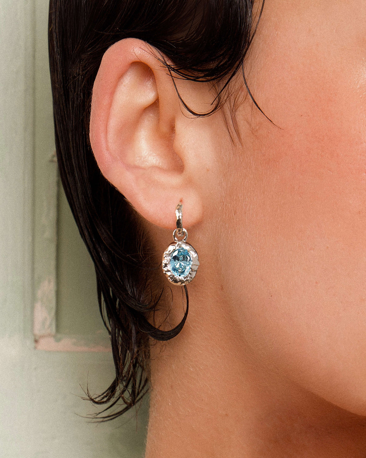 Silver hoop earring with oval light blue birthstone charm. Textured molten frame design.